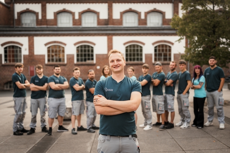 Sie sehen einen jungen sehr aktraktiven Handwerker welcher vor seinem 13-köpfigen Team steht. Alle wirken freundlich und kompetent. Jeder, der dieses Foto sieht, weiß: hier wird hochprofessionelles Handwerk ausgeübt.
