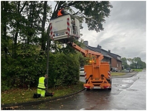 Ein Hubsteiger vor einer Straßenlaterne; im Korb des Hubsteigers sowie am Fuß der Straßenlaterne ist jeweils ein Mann. 