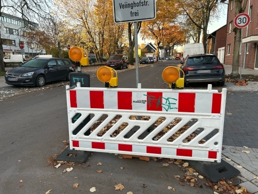 Baustellenzaun an der Hochstraße in Waltrop, im Hintergrund parkende Fahrzeuge, Bäume und Bebauung. 