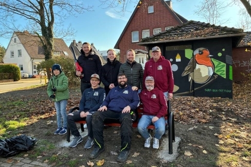 Acht Personen sitzen auf einer Bank bzw. stehen dahinter, im Hintergrund eine Grünfläche mit Laub und ein Trafohäuschen mit einem Bild von einem Rotkehlchen. 