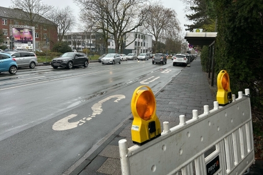 Bushaltestelle gegenüber dem Rathaus, im Vordergrund ein Bauzaun, die Bushaltestelle, im Hintergrund die Straße, Bäume und Gebäude des Rathauses. 