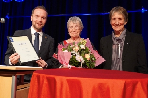 Drei Personen stehen auf einer Bühne, eine hält einen Blumenstrauß, einer eine Urkunde.