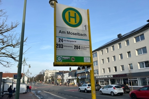 Ein Bushaltestellen-Zeichen, im Hintergrund die Straße Am Moselbach, Autos und Gebäude.  
