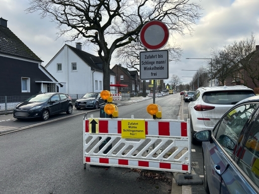 Eine Baustellen-Absperrung, ein Durchfahrt-verboten-Schild, Schild "Zufahrt bis Schlingemann / Winkelheide frei", parkende Autos, eine Straße, Bäume, Häuser.  