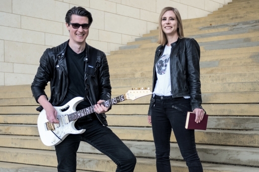 Eine Frau und ein Mann stehen auf einer Treppe, der Mann trägt eine Sonnenbrille und hat ein Instrument in der Hand. 