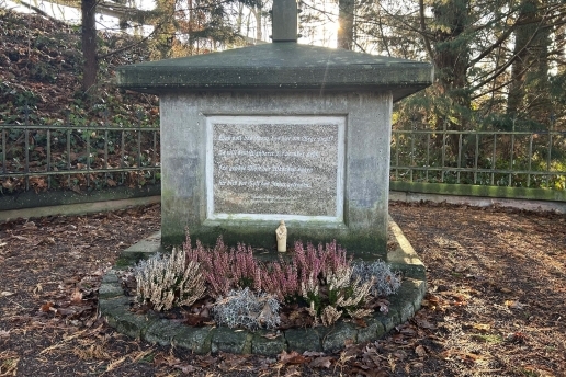 Der Stein-Sockel des Kreuzes Rumsmühle mit einer Inschrift, Bäume im Hintergrund.  