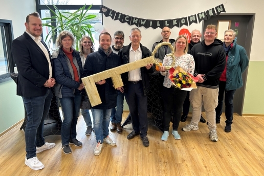 Eine Gruppe von Personen in einem Raum der neuen DRK-Kita, sie halten einen Blumenstrauß und einen überdimensionalen Schlüssel, über ihnen hängt eine schwarze Girlande mit weißer Schrift: herzlich willkommen. 