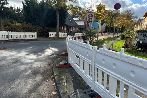 Baustellen-Absperrungen, im Hintergrund eine Straße mit Häusern, Vorgärten, Straßenschildern, Bäumen.