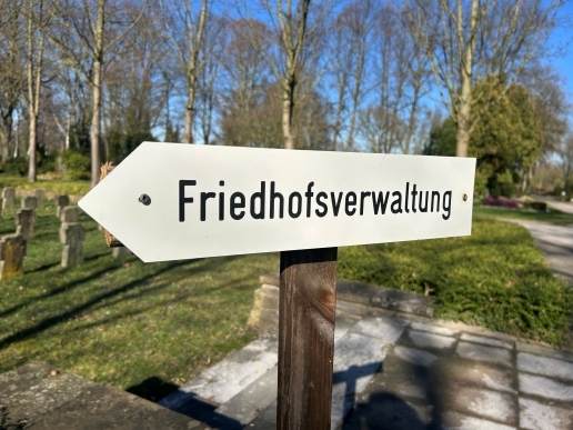 Schild mit der Aufschrift Friedhofsverwaltung, im Hintergrund Bäume, Grabsteine und Grünanlagen des Friedhofs.