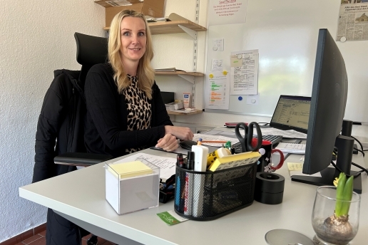 Eine Frau sitzt an einem Schreibtisch, auf dem Schreibtisch-Utensilien und ein Monitor stehen.