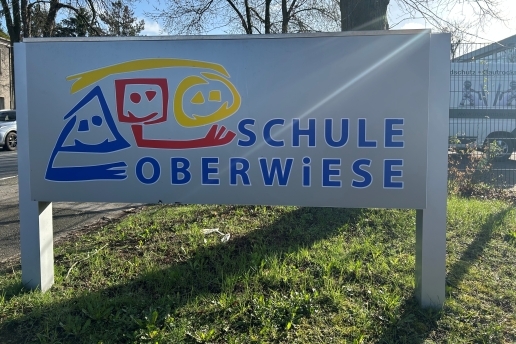 Das Schild Schule Oberwiese in blauer Schrift mit rot-gelb-blauem Logo, das verschiedenförmige Köpfe symbolisiert.