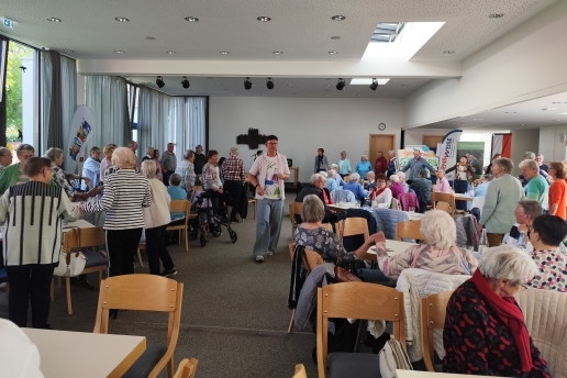 Mehrere Menschen sitzen und stehen an Tischen in einem Saal, teilweise tanzen sie.