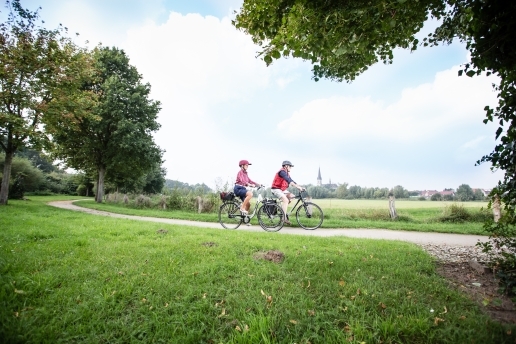 Zwei Fahrradfahrer, im Hintergrund ein Dorf, zudem Bäume und ein Radweg.
