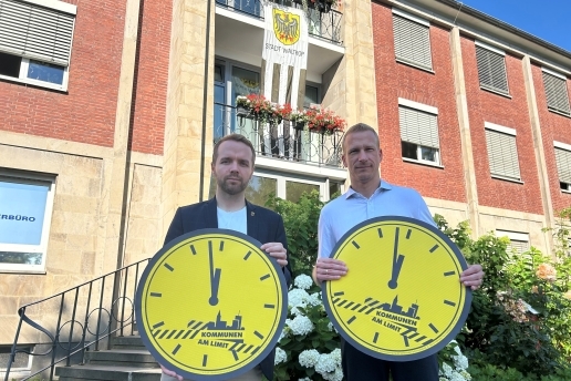 Waltrops Bürgermeister und der Kämmerer stehen mit symbolischen Uhren, die kurz vor 12 Uhr zeigen, vor dem Rathaus.