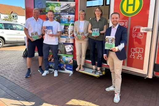 Fünf Personen stehen vor einem roten Fahrzeug und halten VHS-Programme vor sich. 