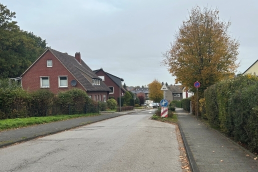 Eine Straße, eine Verkehrsinsel, Bäume, Hecken und Häuser. 