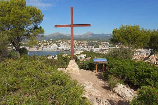 Blick auf eine Bucht auf Mallorca, im Vordergrund ein Kreuz, im Hintergrund Häuser. 
