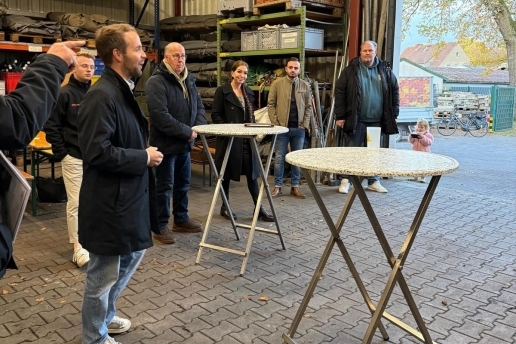 Mehrere Personen stehen rund um zwei Stehtische, im Hintergrund Regale mit Gegenständen. 