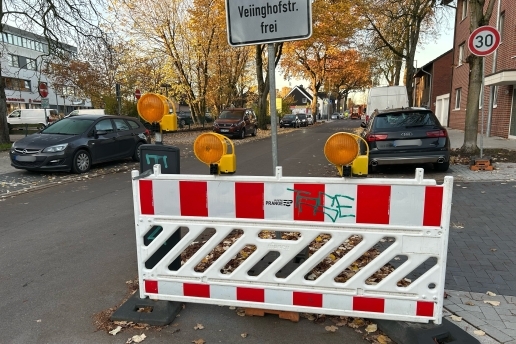 Baustellenzaun an der Hochstraße in Waltrop, im Hintergrund parkende Fahrzeuge, Bäume und Bebauung. 