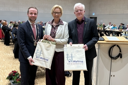 Drei Personen stehen in der Stadthalle: Bürgermeister Marcel Mittelbach (links), Ute Kölker (Mitte) und Klaus Beie. Sie halten Taschen mit Waltrop-Logo in der Hand.