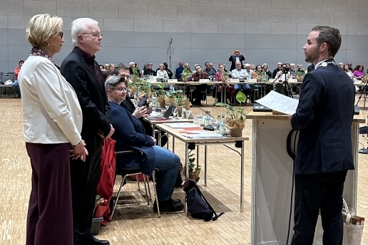 Drei Personen stehen während der Ratssitzung am Rednerpult; im Hintergrund Mitglieder des Rats der Stadt Waltrop und Zuschauende in der Stadthalle. 