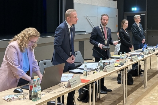 Fünf Personen stehen hinter Tischen mit Redner-Mikrofonen. 