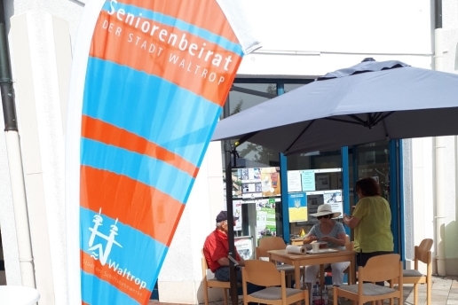 Beachflag des Seniorenbeirats in Orange und Blau mit der Aufschrift Seniorenbeirat der Stadt Waltrop und Logo der Stadt Waltrop; im Hintergrund zwei Personen an einem Tisch, eine stehende Person unter einem Sonnenschirm.   