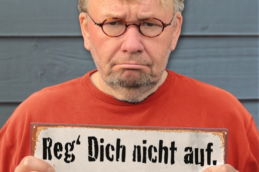 Bernd Stelter in orangefarbenem Pulli blickt grimmig und hält ein Schild "Reg dich nicht auf" in den Händen. 