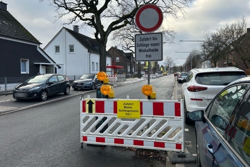 Eine Baustellen-Absperrung, ein Durchfahrt-verboten-Schild, Schild "Zufahrt bis Schlingemann / Winkelheide frei", parkende Autos, eine Straße, Bäume, Häuser.  