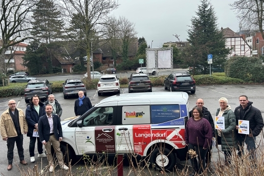 Neun Personen stehen um ein weißes Fahrzeug mit Werbebeflockung herum, im Hintergrund parkende Autos. 