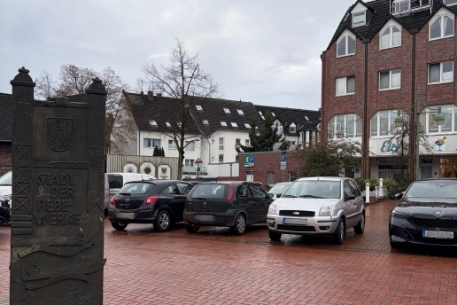 Blick auf den Herne-Bay-Platz mit parkenden Autos, im Vordergrund eine Stele, die auf die Städtepartnerschaft mit Herne Bay hinweist, im Hintergrund Häuser. 