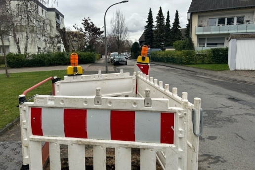 Baustellenabsperrung auf einer Straße, im Hintergrund Mehrfamilienhäuser, parkende Autos, Bäume, Grünflächen. 