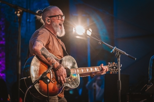 Ein Musiker mit einer Gitarre steht im Scheinwerferlicht auf einer Bühne vor einem Mikrofon.