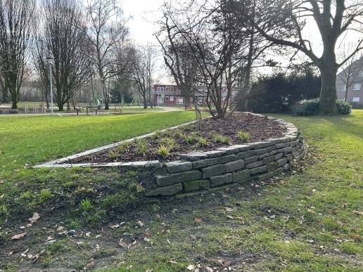 Ein mit Steinen eingefasstes Beet im Stutenteichpark, drum herum Bäume und Rasenflächen sowie eine Wohnanlage.