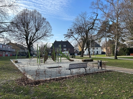 Blick auf den Spielplatz mit Sitzgelegenheit im Stutenteichpark, im Hintergrund Bäume und Rasenflächen.  