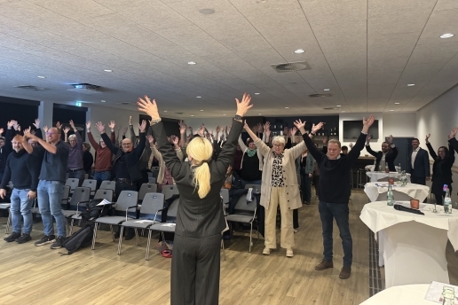 Aktive Pause bei der Auftaktveranstaltung Macherstadt: Menschen stehen in Stuhlreihen und haben die Arme hochgenommen. 