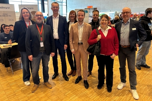 Acht Personen stehen in der Stadthalle, im Hintergrund Messestände und weitere Personen. 