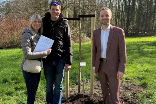 Drei Personen stehen um einen jungen Baum herum, im Hintergrund Wiesenfläche und Bäume. 