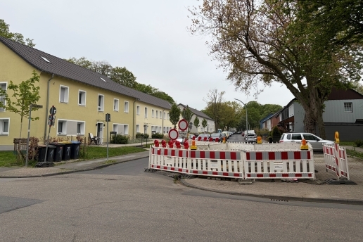 Bauzäune an einer Straßen-Einmündung, Häuser und Bäume im Hintergrund.