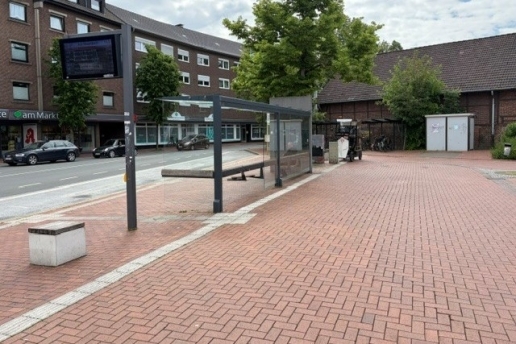 Gepflasterter Platz, eine Bushaltestelle, eine Straße und Häuser im Hintergrund. 