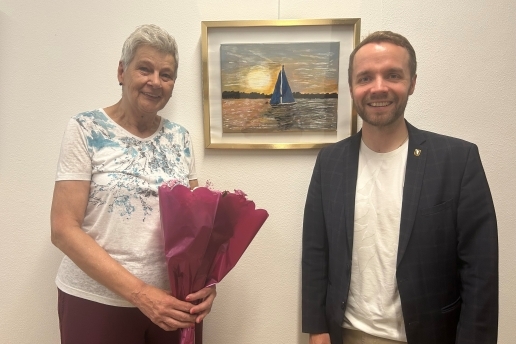 Bärbel Voigt steht mit einem Strauß Blumen neben Bürgermeister Marcel Mittelbach vor einem Bild, das ein Segelschiff und einen Sonnenuntergang zeigt.  