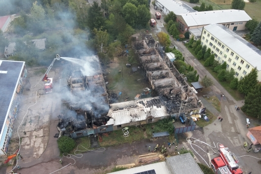 Luftaufnahme eines Gebäude-Ensembles, aus dem Rauch quillt; große Teile des Gebäudes sind von einem Brand zerstört.. 
