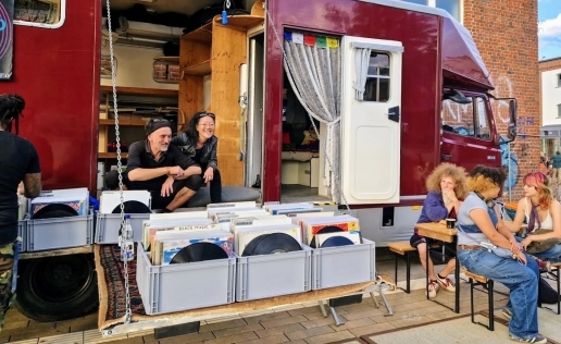 Personen sitzen vor einem Camper-Mobil mit Schallplatten-Kisten.