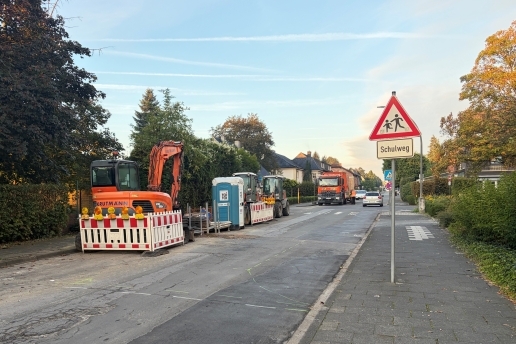Eine Straße, links Bauzäune, eine mobile Toilette, mehrere Baufahrzeuge, rechts ein Schild "Schulweg". 