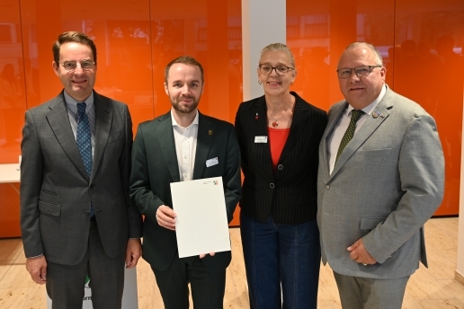 Vier Personen stehen vor einer orangefarbenen Wand, eine Person hält einen Bescheid in der Hand. 