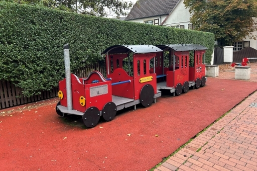 Eine rote Spielzeug-Lok samt Anhänger auf rotem Fallschutz vor einer Hecke.