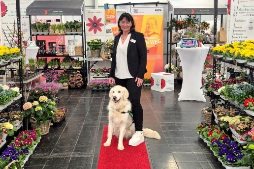 Eine Frau, zu deren Füßen ein Hund mit hellem Fell sitzt, steht in einem Floristik-Geschäft. 